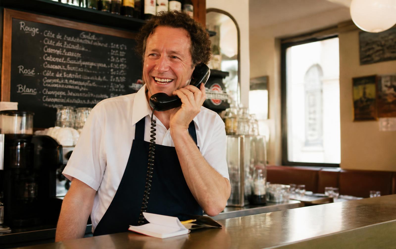 Leo lacht am Tresen mit dem Telefonhörer am Ohr, daneben das Reservierungsbuch