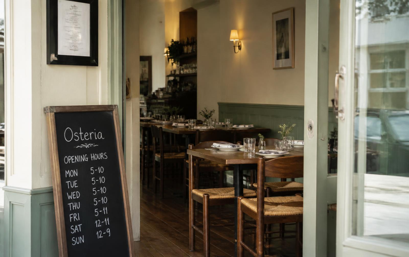 Blick durch die offene Tür in den Gastraum, davor eine Tafel mit den Öffnungszeiten